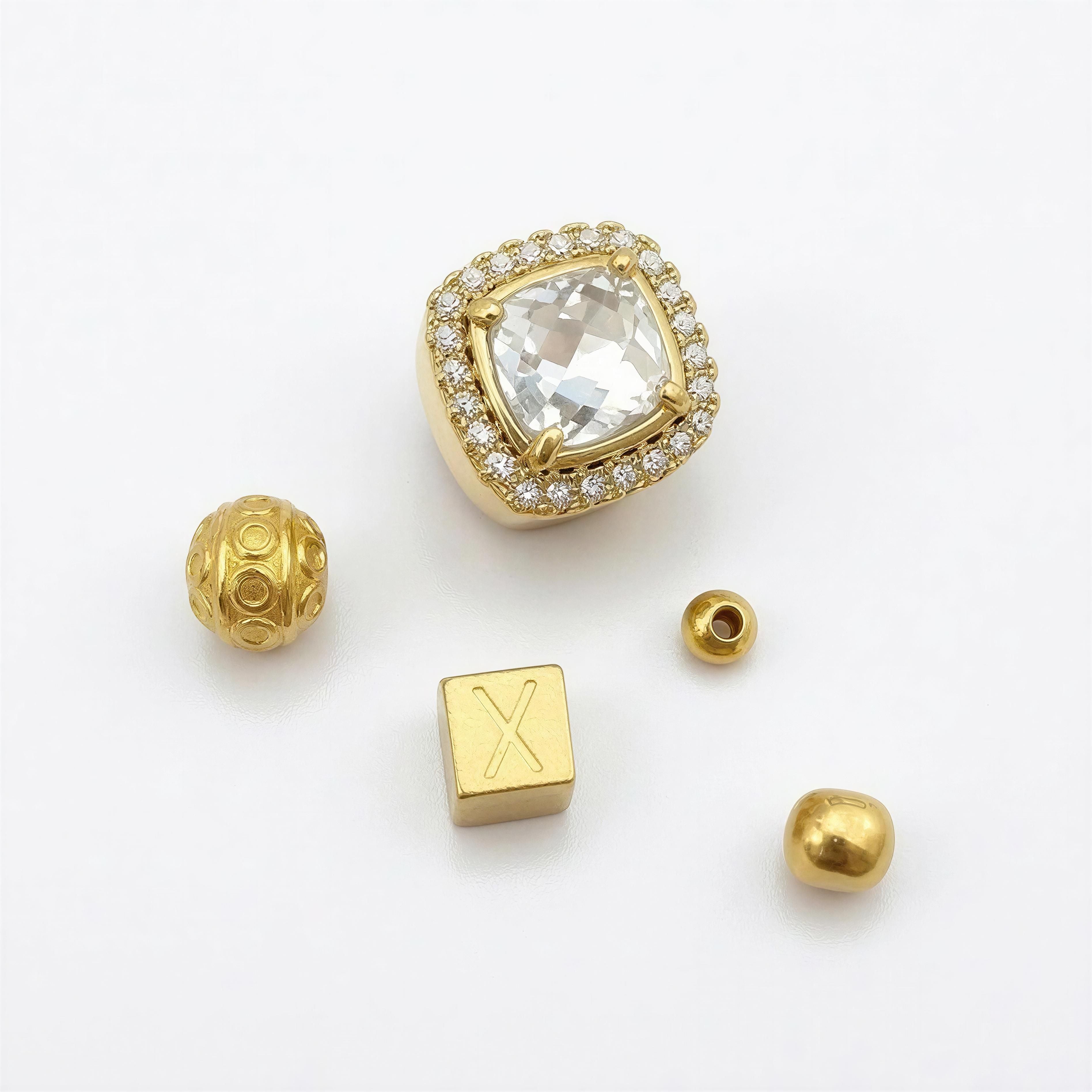 Close-up of five luxury beads including a gold and crystal centerpiece surrounded by small diamonds, a textured gold bead, a smooth gold bead, a hollow gold cylinder bead, and a gold cube bead engraved with an X, part of the XX ÉLÉGANCE LIMITED EDITION collection.