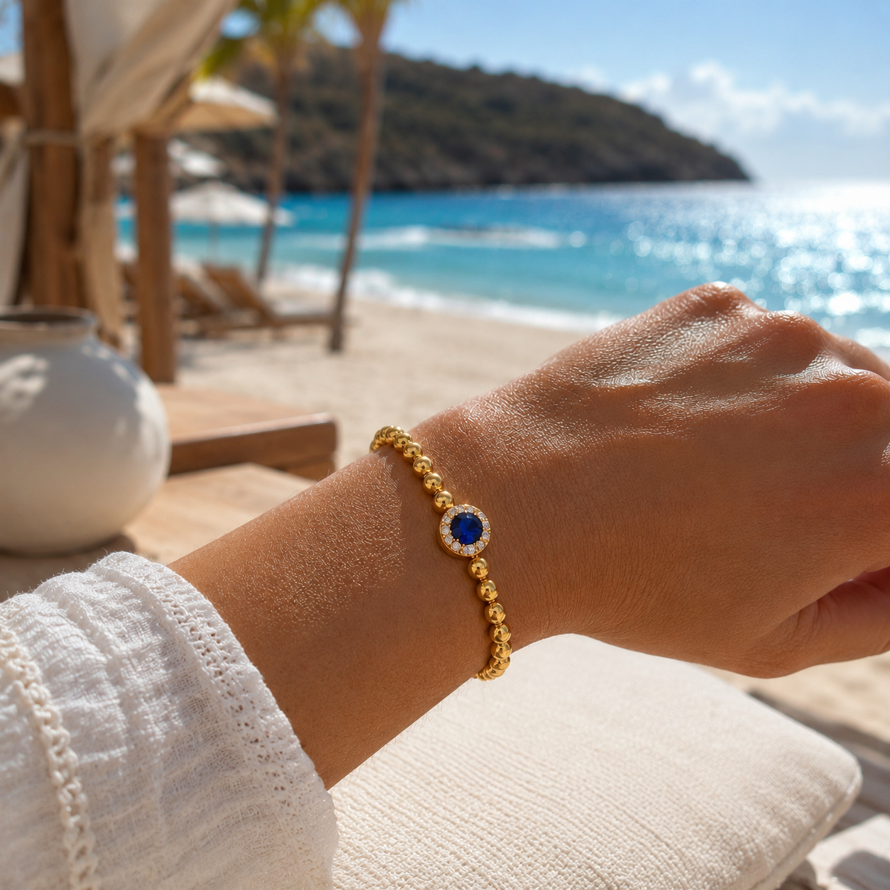 Close-up of a wrist wearing the XX ÉLÉGANCE - AZURA ROYALE gold beaded bracelet with a deep blue gemstone, set against a sunlit beach and ocean background.