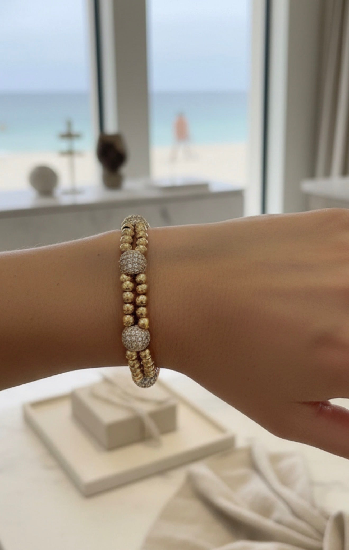 Close-up of a wrist wearing the XX Élégance – Oro Celestia bracelet featuring gold beads and sparkling pavé spheres, set against a soft beach view background.