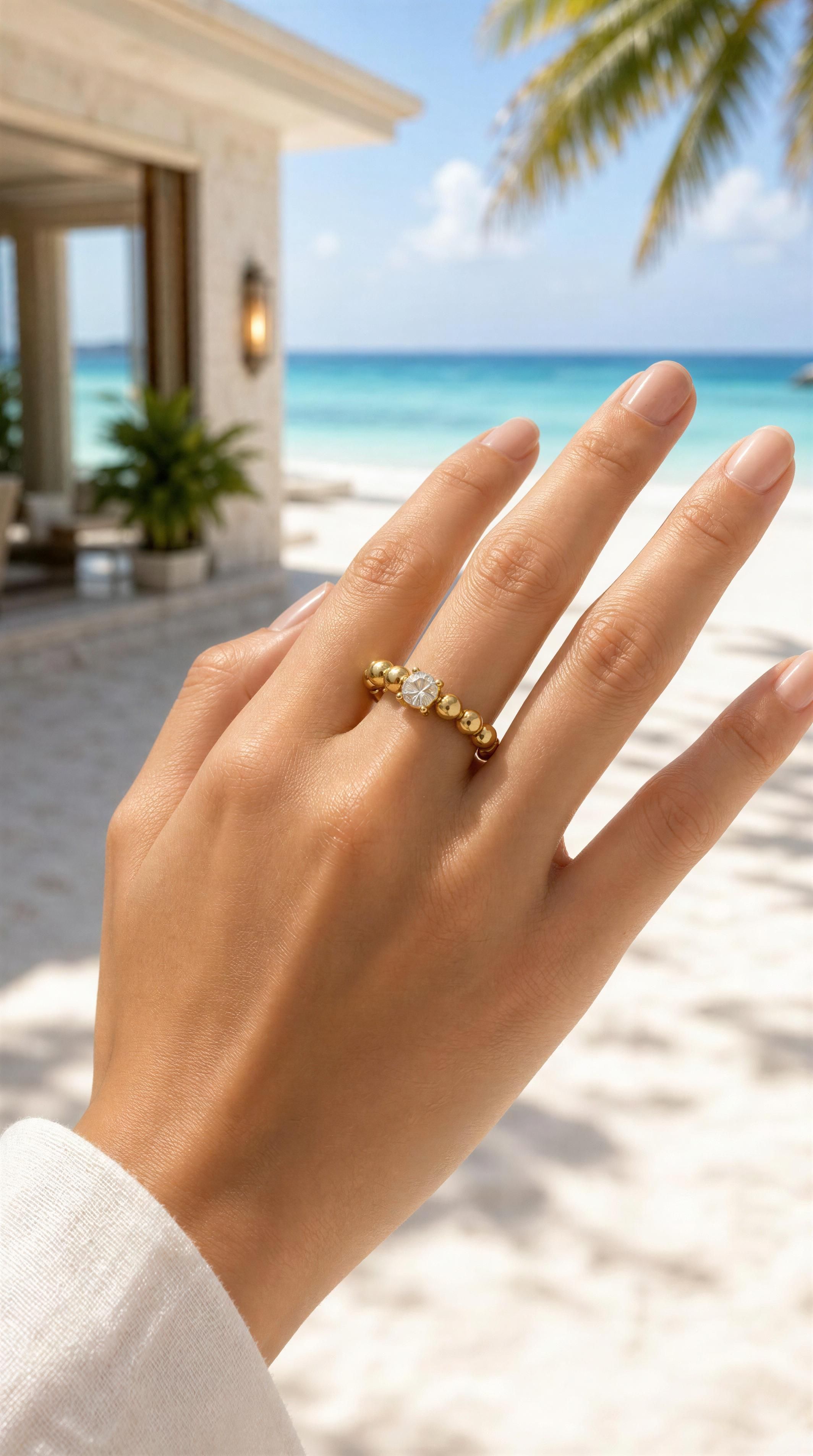 Hand wearing a RING - LUCENT ΓTOILE RING featuring gold beads and a central clear gemstone, with a serene beach and tropical palm background.