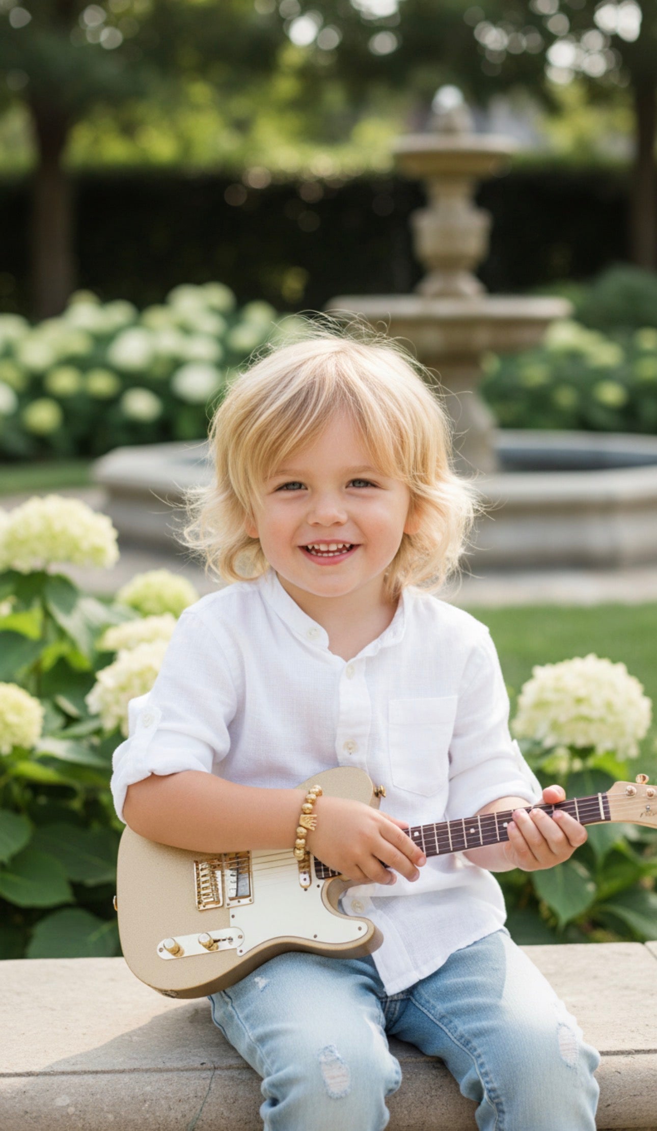 Young child smiling outdoors wearing The Little Icons - Manus Ferox bracelet while holding a small electric guitar in a garden setting with soft natural light.