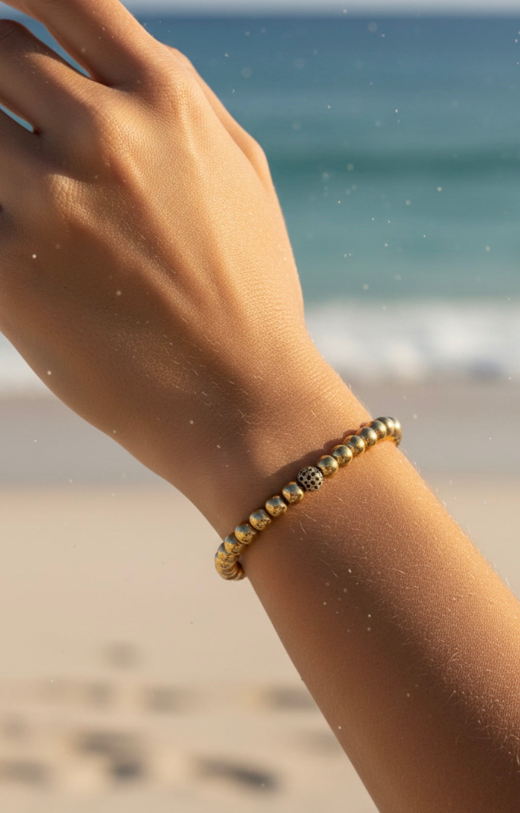Close-up of a wrist wearing a gold-colored beaded bracelet from the Timeless Collection - Essentia Nox, set against a blurred beach background.