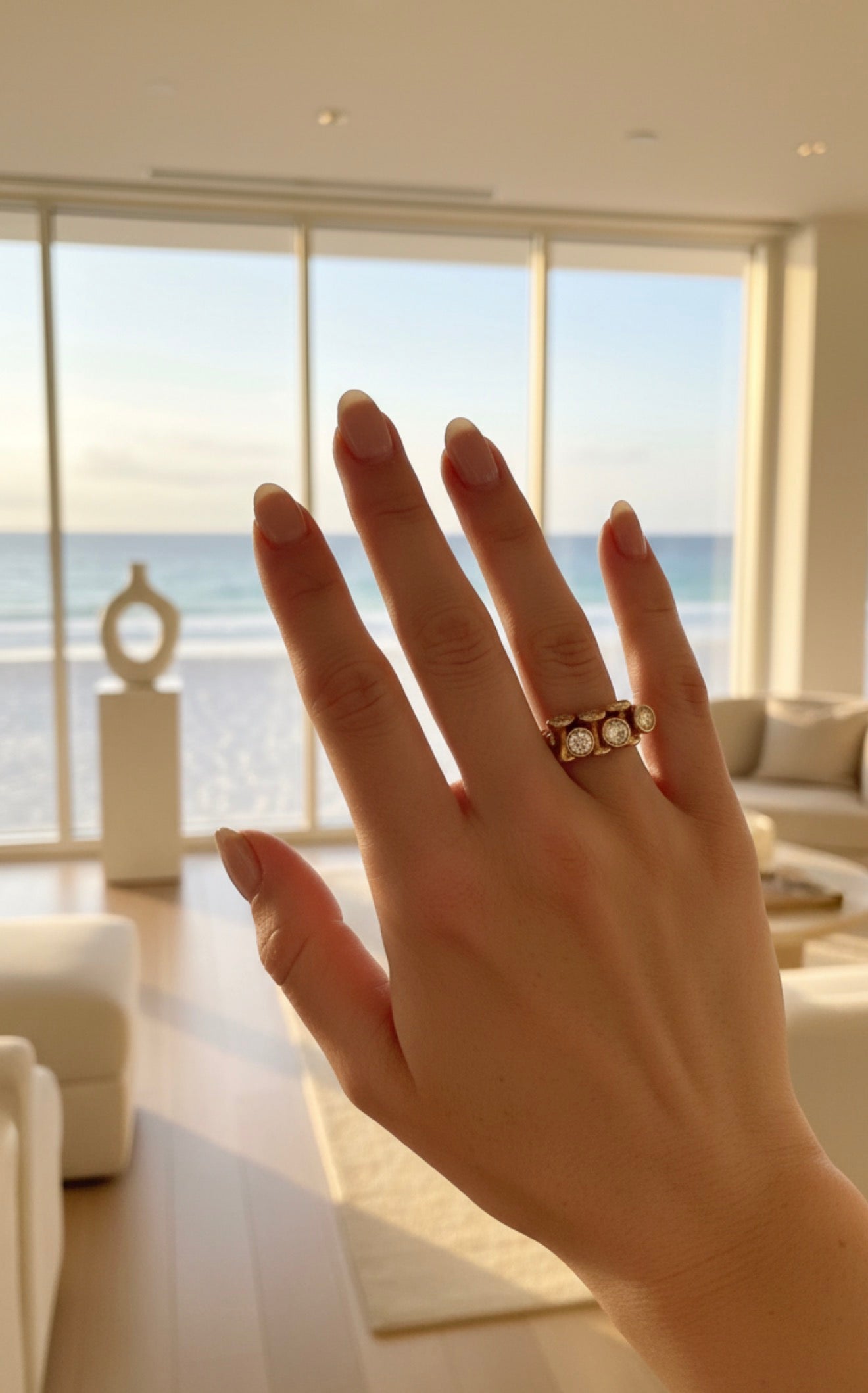 Hand wearing the RING Golden Light featuring multiple round gemstones set in a gold band, with a sunlit modern living room and ocean view in the background.