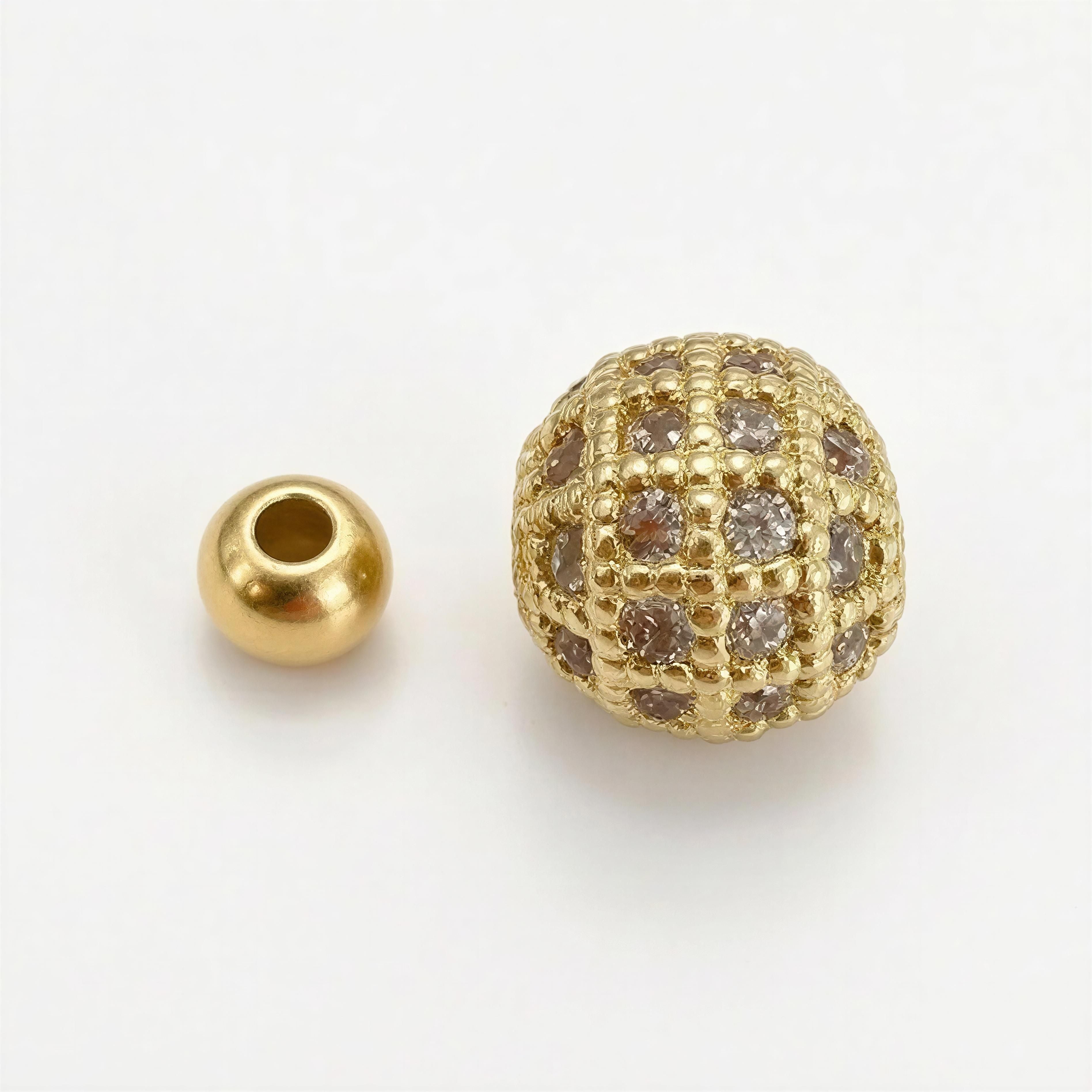 Two gold beads including a smooth spherical bead and an intricately designed textured bead with encrusted stones for the XX Élégance – Oro Celestia handcrafted luxury jewelry collection.