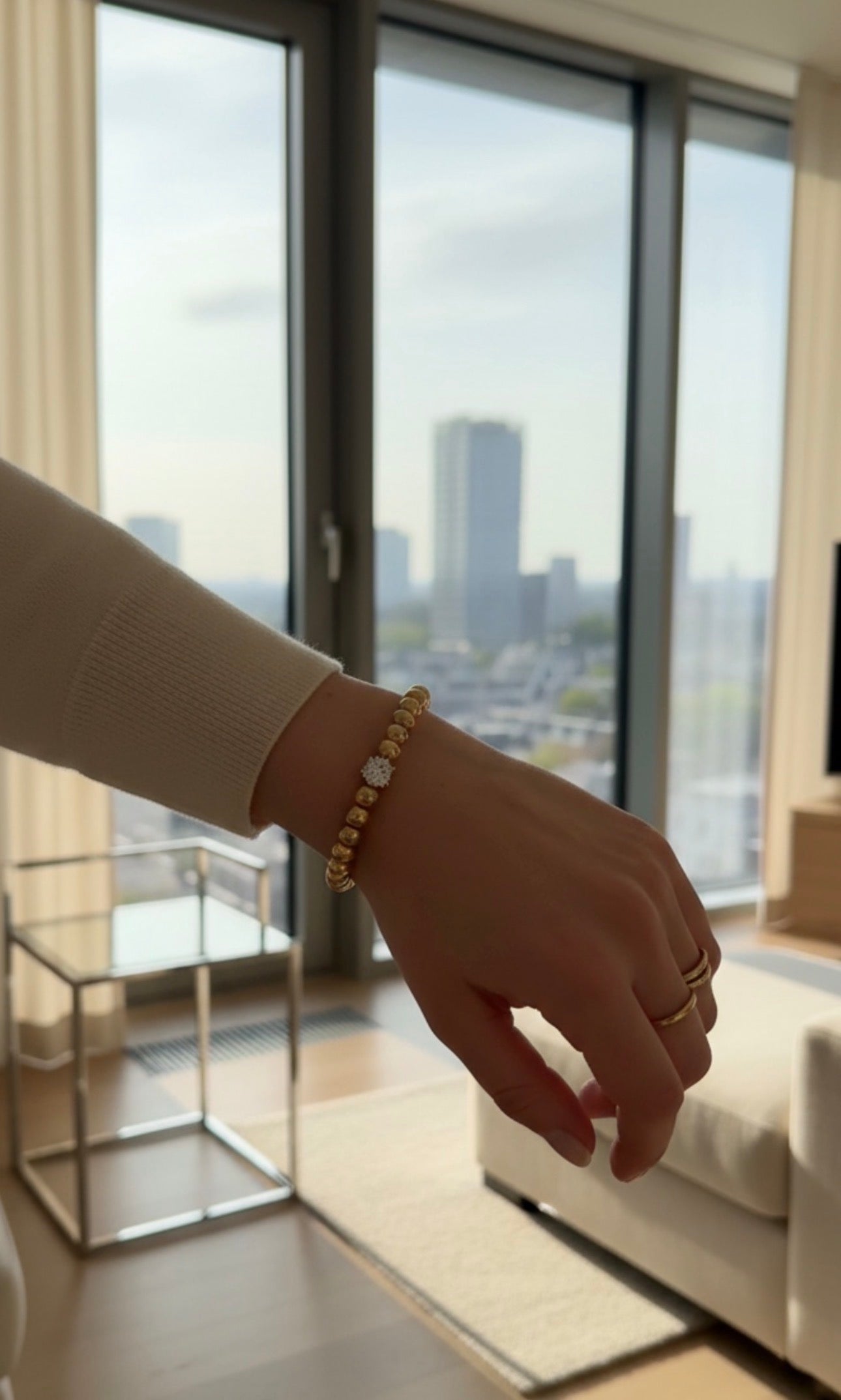 Close-up of a wrist adorned with a gold beaded bracelet featuring a central decorative bead, worn inside a modern living room with city views, showcasing The Timeless Collection – Aurea Claritas.
