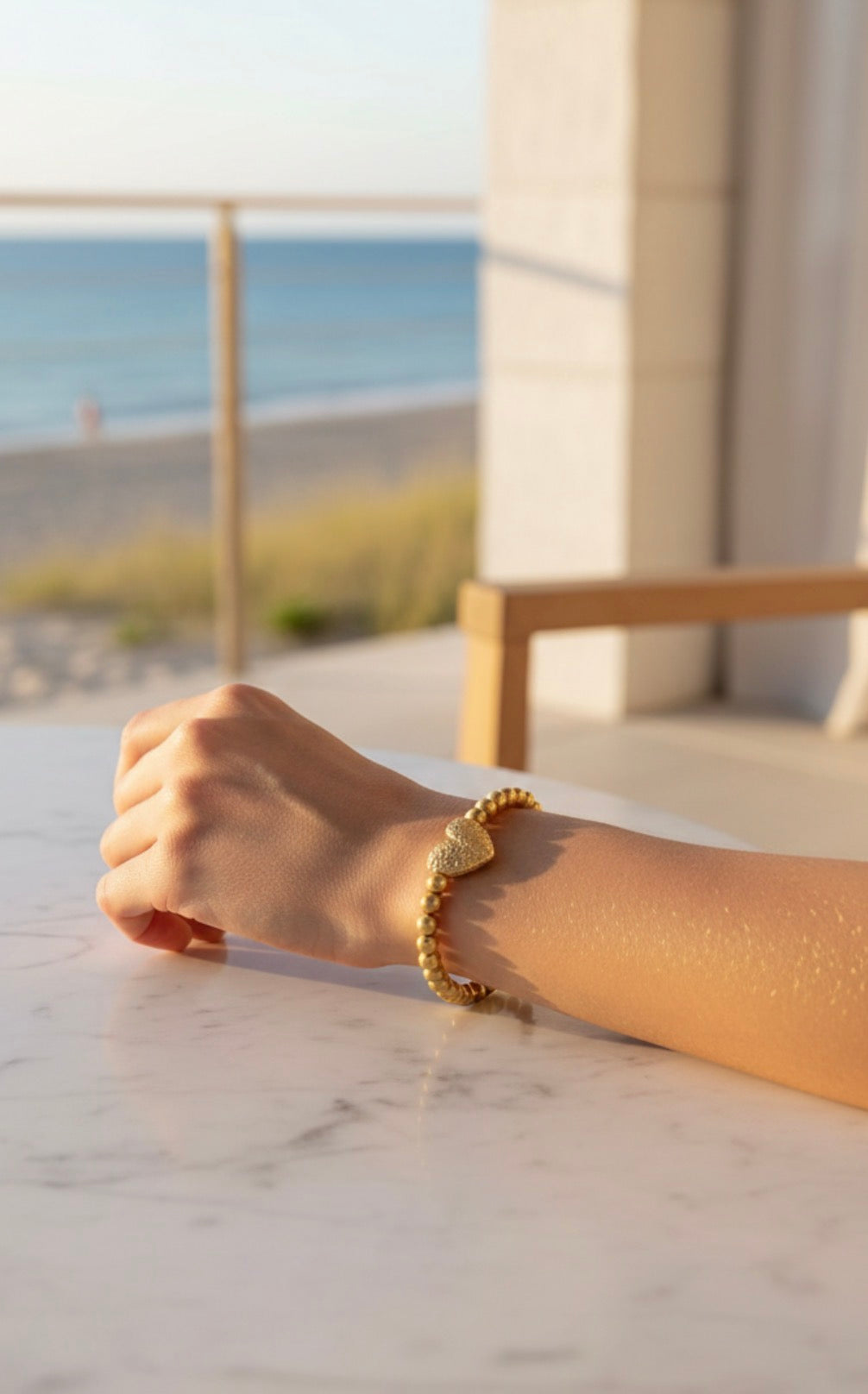 Close-up of a wrist adorned with a gold beaded bracelet featuring a heart-shaped charm, set on a marble table with a serene beach view in the background, showcasing the Timeless Collection - Amor Aurea.