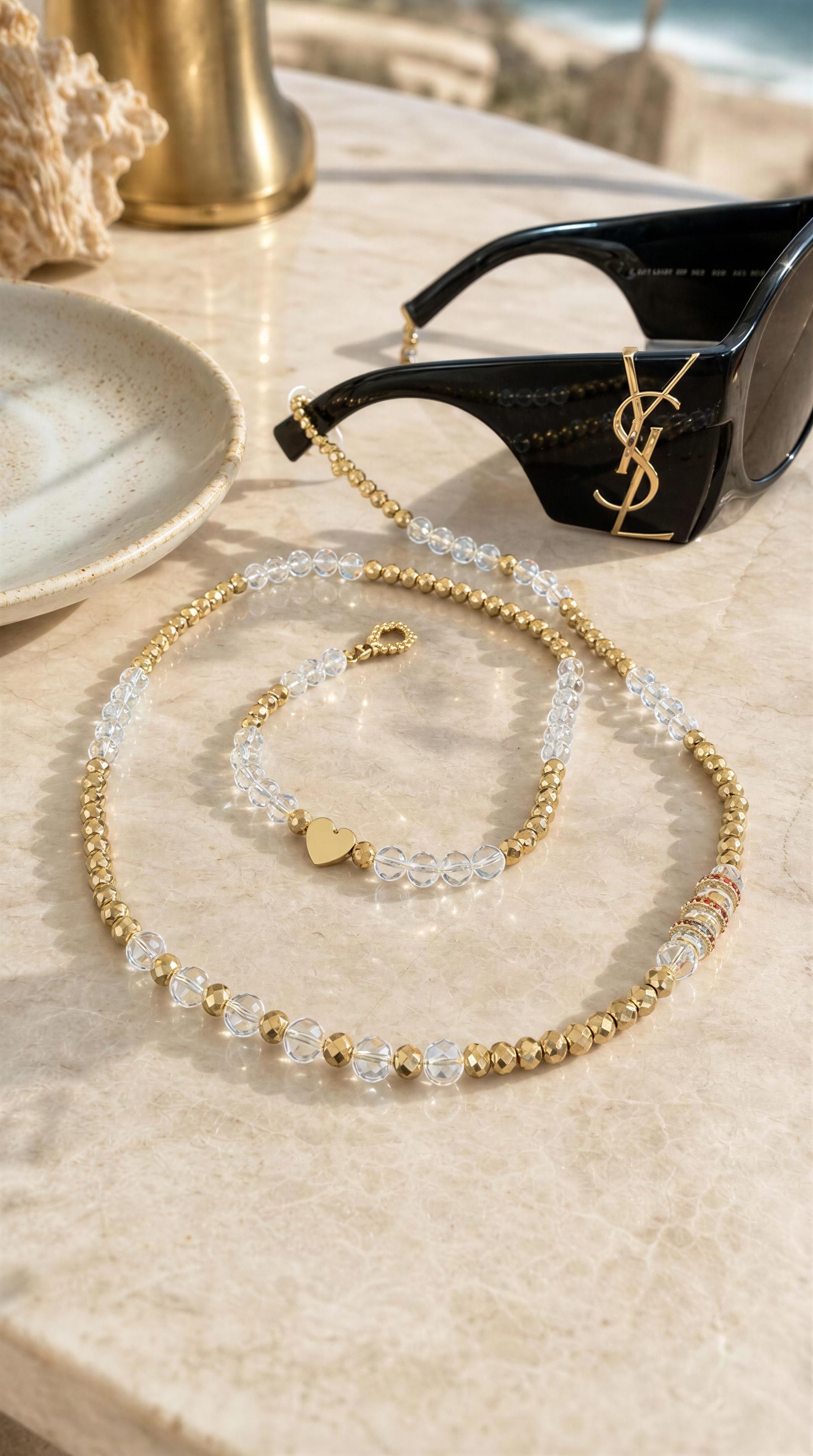 The Riviera Sunglass Chain - Cristalle features alternating clear and gold beads with a delicate heart charm, elegantly draped next to black designer sunglasses on a marble surface.