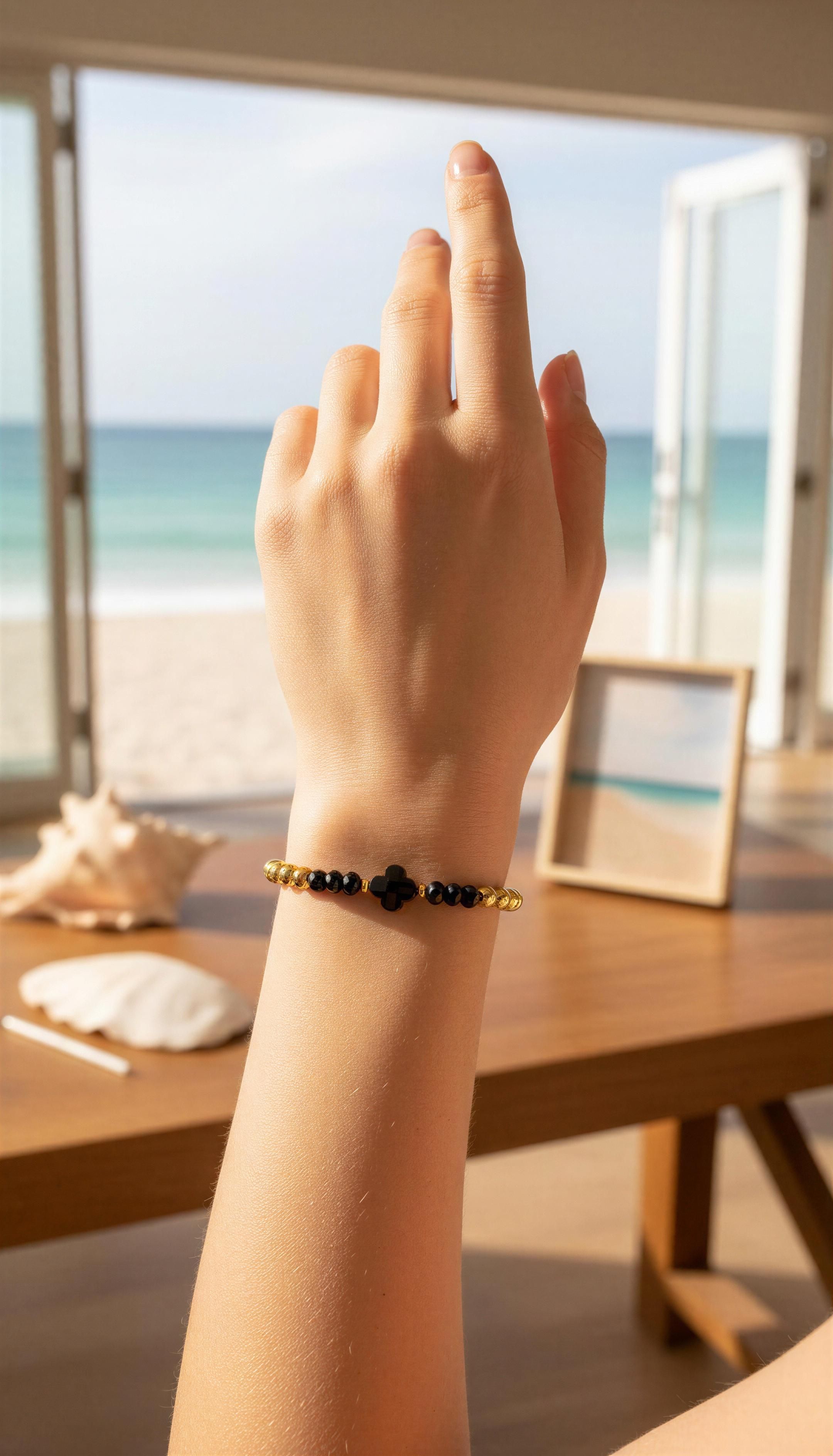Hand wearing XX ÉLÉGANCE - Noir Fortuna bracelet featuring black and gold beads with a black clover charm, set against a bright beachside background.