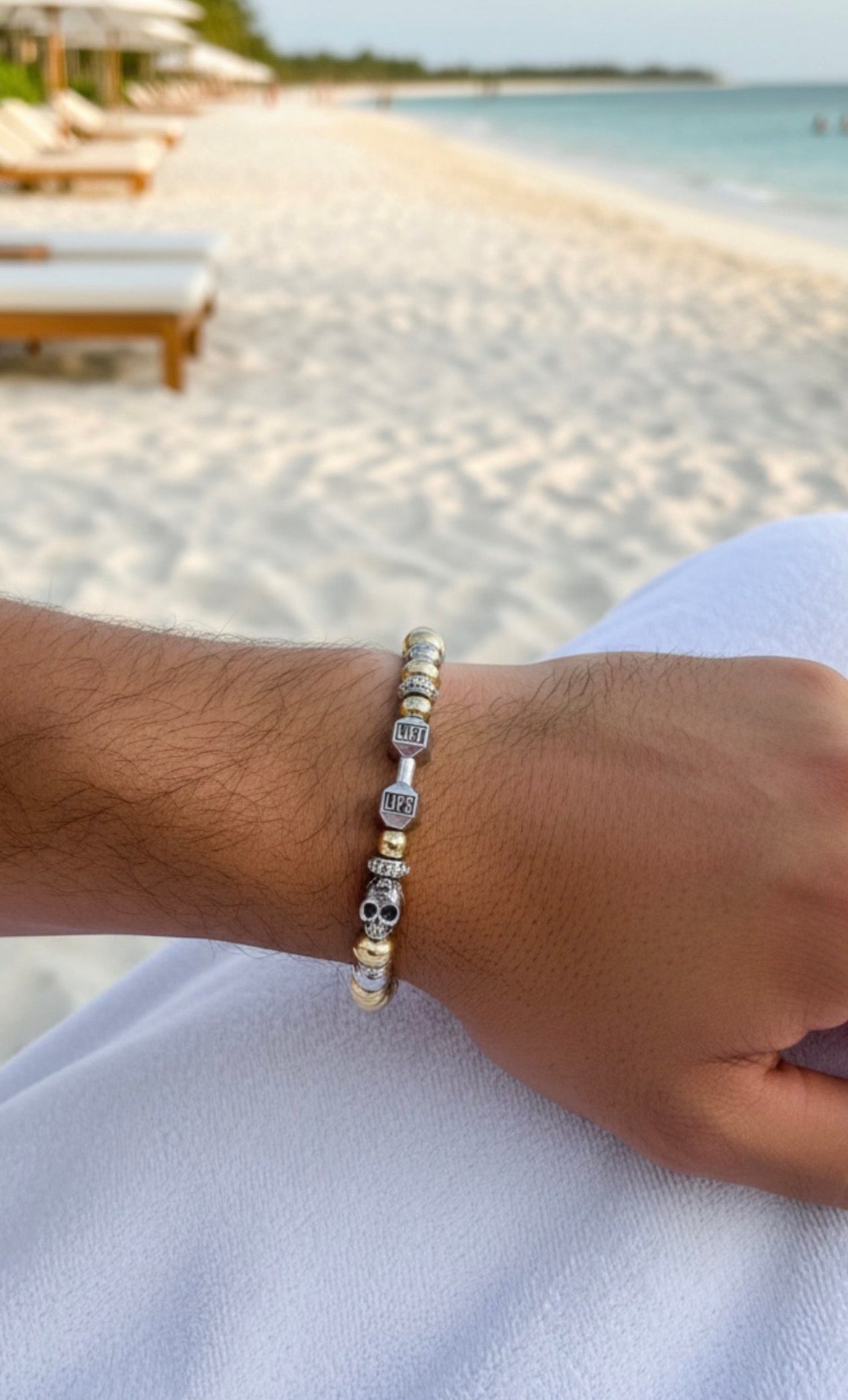 Close-up of a wrist wearing The Iron Legacy Bracelet featuring gold and silver beads with skull and cube charms, set against a serene beach background.