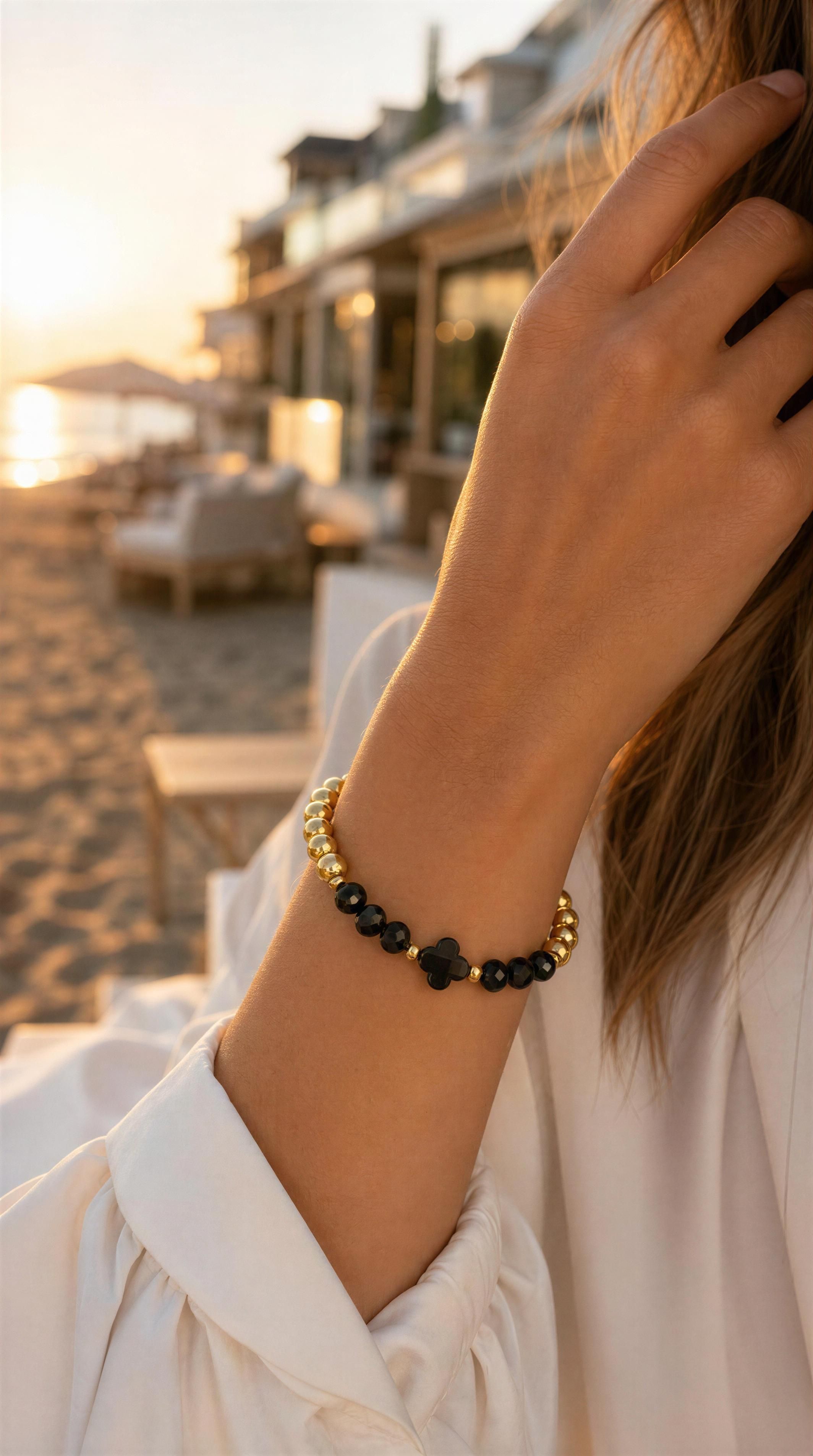 Close-up of a wrist wearing the XX ÉLÉGANCE - Noir Clover bracelet featuring black clover and gold beads, with a serene beach scene at sunset in the background.