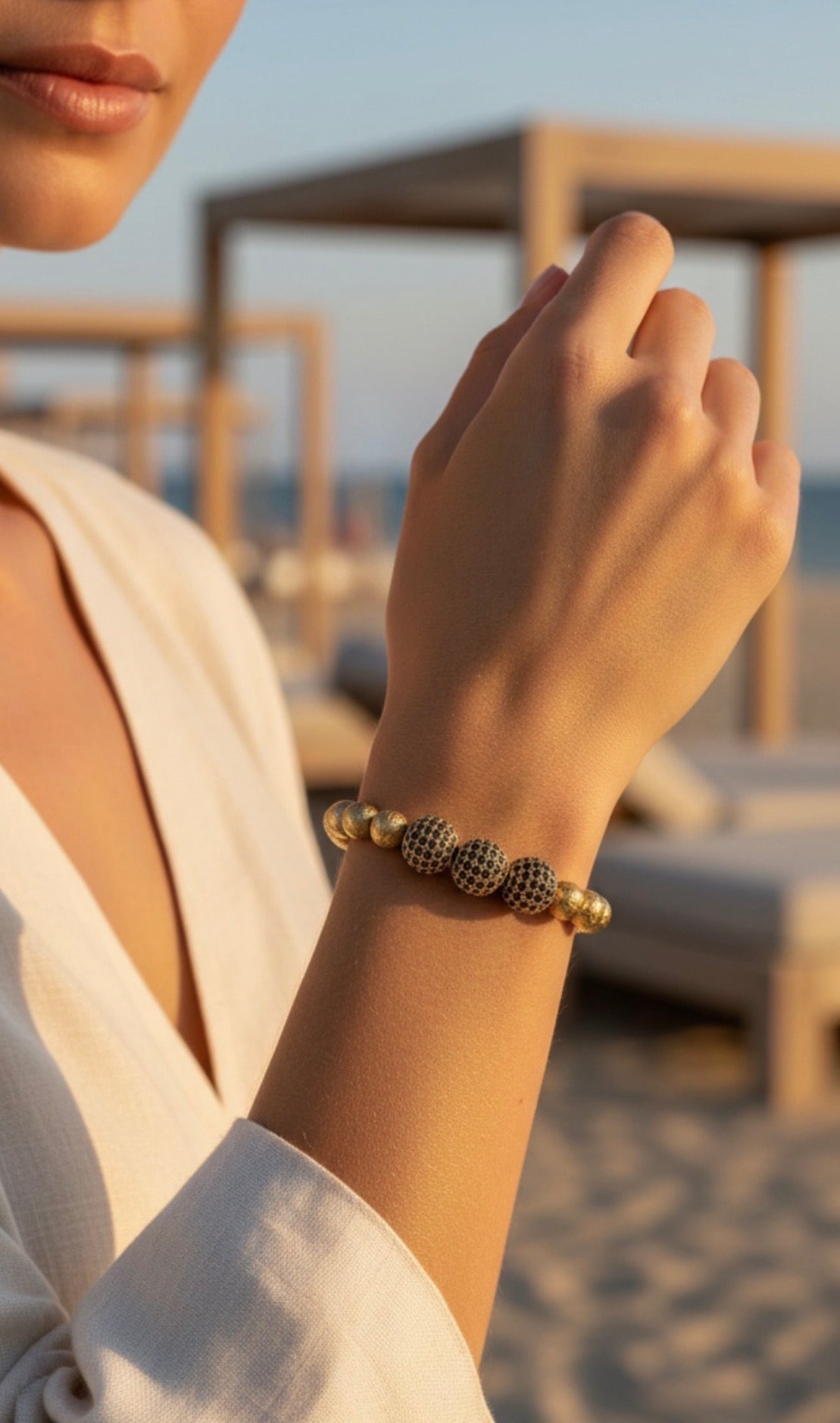 Close-up of a wrist adorned with the Limited Edition- Aurea Nox Imperium bracelet featuring gold beads and textured black accents, set against a serene beach background.