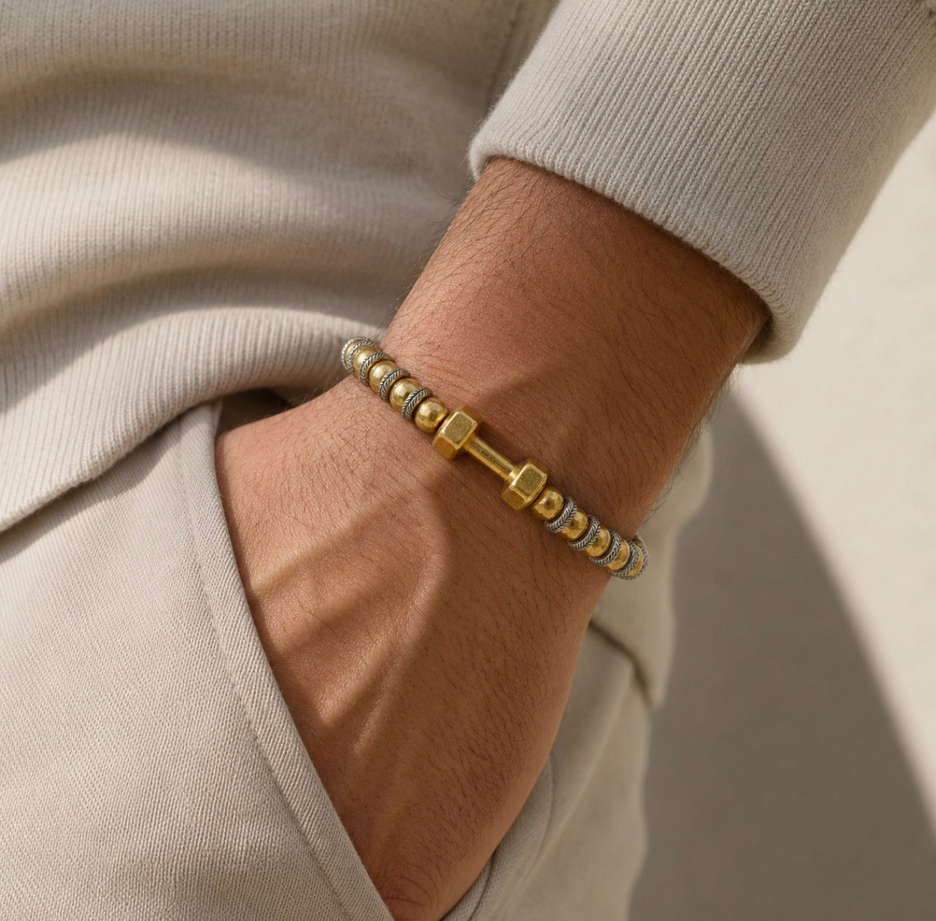 Close-up of a wrist wearing THE X FOR HIM - Driven Balance bracelet featuring gold and silver beads with a central gold hex nut design on a beige outfit background.