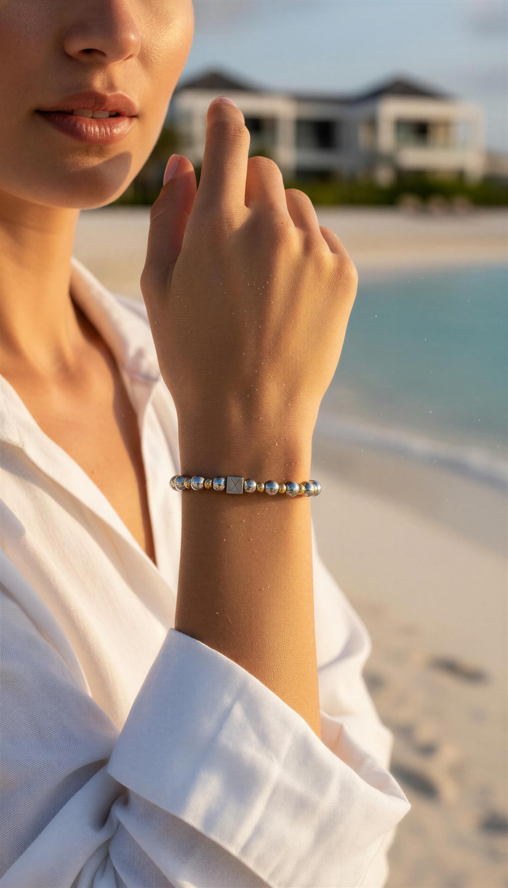 Close-up of a woman's wrist wearing The Essential Collection - 6mm & 4mm gold and silver beaded bracelet, with a blurred beach and modern houses in the background.